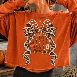 May include: A burnt orange long-sleeve sweatshirt featuring a large central graphic of a bow with a leopard print pattern and white polka dots. Dangling from the bow are beaded strings with charms including a pumpkin, acorn, leaf, sunflower, and mug.