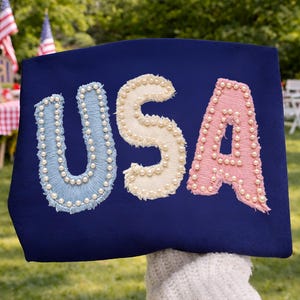 K&ouml;nnte beinhalten: Ein marineblaues Textil mit dem Wort "USA", gestickt in Hellblau, Creme und Rosa. Jeder Buchstabe ist mit kleinen, wei&szlig;en Perlen verziert. Der Hintergrund zeigt eine gr&uuml;ne Au&szlig;enumgebung.