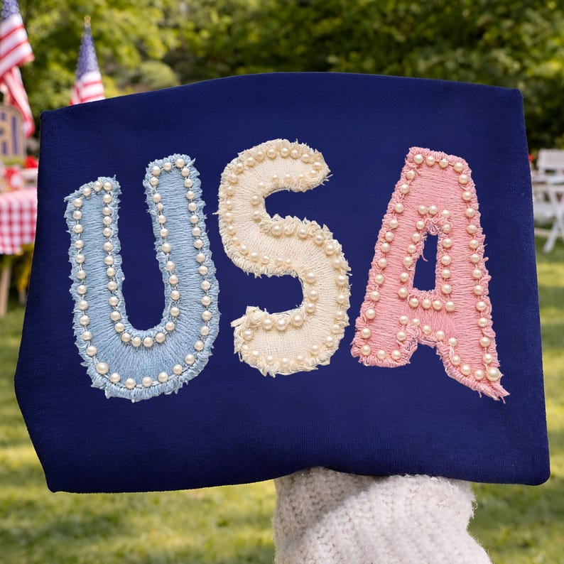 K&ouml;nnte beinhalten: Ein marineblaues Textil mit dem Wort "USA", gestickt in Hellblau, Creme und Rosa. Jeder Buchstabe ist mit kleinen, wei&szlig;en Perlen verziert. Der Hintergrund zeigt eine gr&uuml;ne Au&szlig;enumgebung.