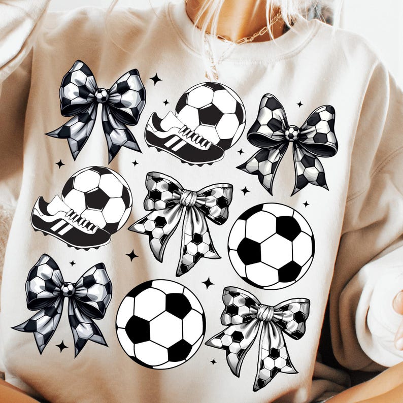 May include: Cream-colored sweatshirt featuring a soccer theme. The design includes soccer balls, soccer cleats, and bows with a soccer ball pattern. Black star accents are scattered around the design.