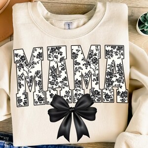 May include: Cream-colored sweatshirt with the word "MAMA" in large, black floral print letters. A large black bow is centered below the text. The sweatshirt has a crew neck and long sleeves.