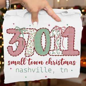 May include: White t-shirt featuring the numbers "3701" in red and green plaid patterns, with a bow on each number. The text "small town christmas" and "nashville, tn" are printed below. The design has a festive holiday theme.