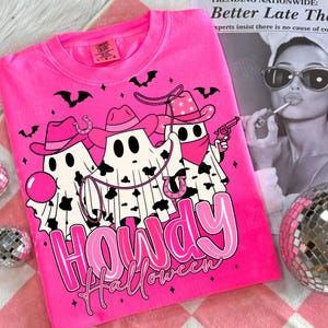 May include: A bright pink sweatshirt featuring a graphic of three ghost cowboys with cowboy hats, bandanas, and a lasso. The text "Howdy Halloween" is displayed in pink. Bats and horseshoes are also featured in the design.