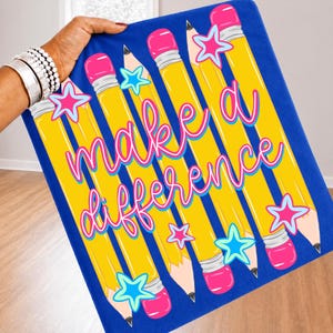 May include: A blue fabric surface featuring a repeating pattern of yellow pencils with pink erasers and light blue star shapes. The text "make a difference" is printed in a cursive pink font across the center of the pencils and stars.