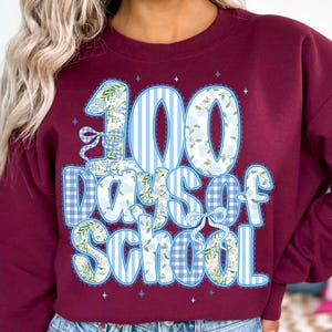 K&ouml;nnte beinhalten: Burgunderfarbenes Sweatshirt mit einer gro&szlig;en Grafik, die 100 Schultage feiert. Das Design zeigt die Zahl "100" in blau-wei&szlig;en Streifen und floralen Mustern, mit den Worten "Days of School" in einer verspielten Schriftart.