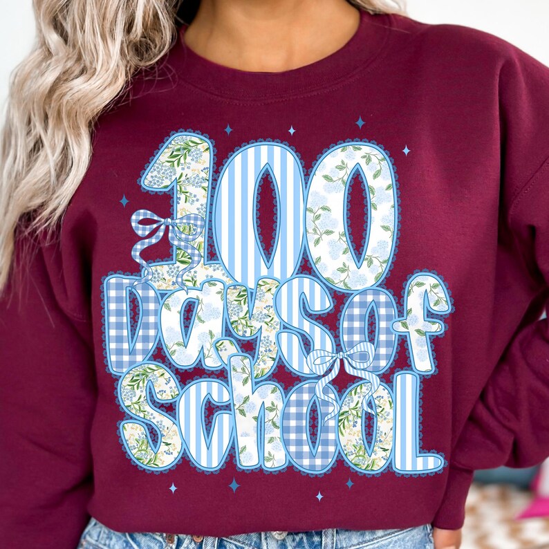 K&ouml;nnte beinhalten: Burgunderfarbenes Sweatshirt mit einer gro&szlig;en Grafik, die 100 Schultage feiert. Das Design zeigt die Zahl "100" in blau-wei&szlig;en Streifen und floralen Mustern, mit den Worten "Days of School" in einer verspielten Schriftart.