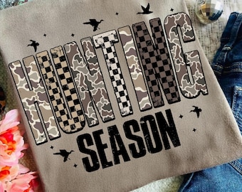 May include: A beige t-shirt featuring the text "HUNTING SEASON". The letters are a mix of camouflage and chequered patterns. The design includes bird and star silhouettes. The shirt is made of a soft material.
