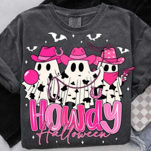 May include: Dark gray t-shirt featuring a Halloween-themed graphic. The design includes three ghost figures wearing pink cowboy hats and bandanas, with a lasso and a toy gun. The text "HOWDY Halloween" is displayed in pink.