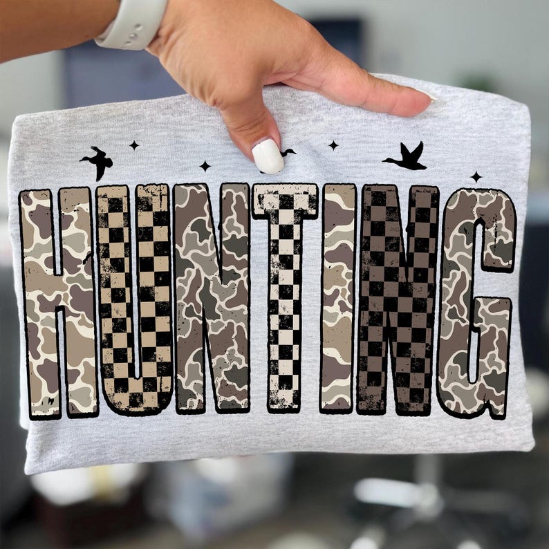 Puede incluir: Una camiseta gris claro con la palabra "HUNTING" en letras grandes y en negrita. Las letras tienen un patr&oacute;n de camuflaje y un patr&oacute;n de cuadros. Encima de la palabra hay tres siluetas de p&aacute;jaros negros y peque&ntilde;as estrellas.