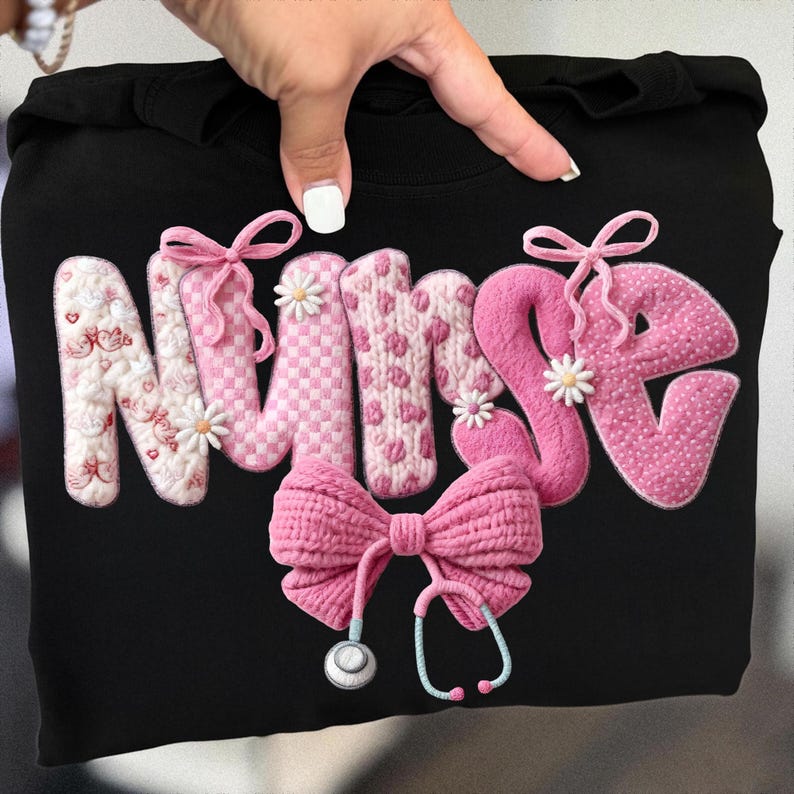 May include: Black sweatshirt with the word "NURSE" in pink, textured letters. The "N" is decorated with hearts and flowers, the "U" and "R" have a checkered pattern, and the "S" and "E" have a polka dot pattern. A pink bow and stethoscope complete the design.