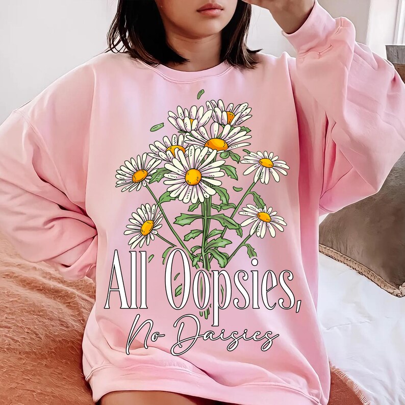 K&ouml;nnte beinhalten: Hellrosa Sweatshirt mit einem G&auml;nsebl&uuml;mchen-Bouquet-Design. Die G&auml;nsebl&uuml;mchen haben wei&szlig;e Bl&uuml;tenbl&auml;tter und gelbe Zentren. Der Text "All Oopsies, No Daisies" ist darunter gedruckt. Das Sweatshirt hat eine hellrosa Farbe.