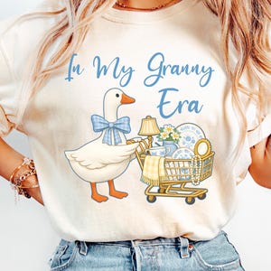Puede incluir: Sudadera color crema con un ganso de dibujos animados con un lazo azul empujando un carrito de compras lleno de art&iacute;culos vintage. El texto "In My Granny Era" est&aacute; escrito en azul sobre el ganso.