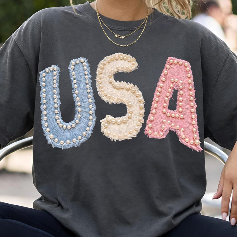 K&ouml;nnte beinhalten: Dunkelgraues Sweatshirt mit dem Wort "USA" in gro&szlig;en, gestickten Buchstaben. Die Buchstaben sind in Hellblau, Creme und Rosa gehalten, jeweils mit kleinen, wei&szlig;en Perlen umrandet. Das Sweatshirt hat einen Vintage-Look.