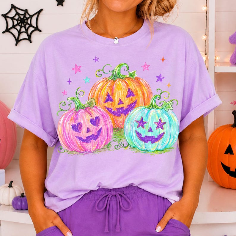 May include: A light purple short-sleeve t-shirt featuring a graphic print of three jack-o'-lanterns in pink, orange, and teal. The pumpkins have heart and star-shaped eyes, and the design is accented with small colorful stars and green vines.