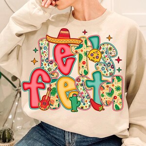 May include: A beige sweatshirt with the words "Let's Fiesta" in colorful, floral-patterned letters. The design includes a sombrero, guitar, cactus, skull, chili pepper, and stars, celebrating a festive theme.
