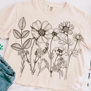 Może przedstawiać: Jasnor&oacute;żowy t-shirt z czarnym rysunkiem linii bukietu kwiat&oacute;w. Kwiaty obejmują duży kwiat z wieloma płatkami, mniejszy kwiat z pięcioma płatkami i kwiat z gwiaździstym środkiem.