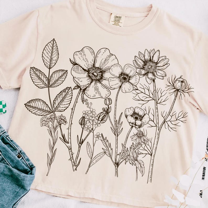 Może przedstawiać: Jasnor&oacute;żowy t-shirt z czarnym rysunkiem linii bukietu kwiat&oacute;w. Kwiaty obejmują duży kwiat z wieloma płatkami, mniejszy kwiat z pięcioma płatkami i kwiat z gwiaździstym środkiem.