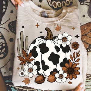 May include: A beige sweatshirt featuring a white pumpkin with black spots, surrounded by brown flowers, leaves, and a cactus. The design includes white flowers, acorns, and star accents, creating a fall-themed graphic.