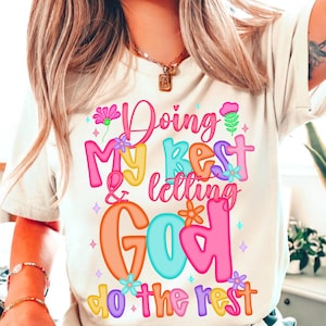 May include: White t-shirt with a colorful graphic that says "Doing my best & letting God do the rest". The graphic features flowers, stars, and a heart.
