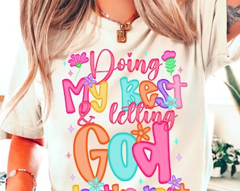May include: Cream-colored t-shirt with a colorful graphic that reads "Doing My Best & Letting God do the rest." The text is in bubble letters with floral and star accents. The shirt is a casual wear item.