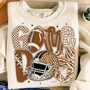 Puede incluir: Sudadera color crema con un gráfico de "GAME DAY". El diseño presenta un balón de fútbol, un casco y letras con varios patrones, incluyendo cuadros, leopardo y estampados de cebra, todo en tonos marrones y beige. Acentos de diamantes negros completan el diseño.