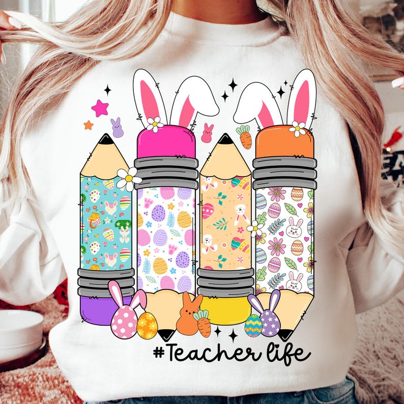 Op de afbeelding: Wit sweatshirt met een afbeelding van drie potloden met konijnenoren. De potloden zijn versierd met paasmotieven, waaronder eieren, konijnen en bloemen. De tekst "#Teacher life" is onder de potloden gedrukt.