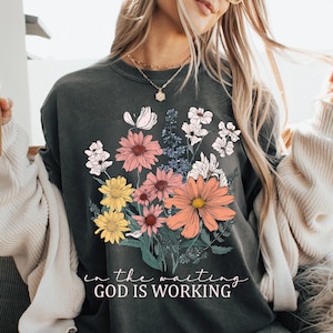 May include: A dark grey t-shirt with a floral design and the text "in the waiting GOD IS WORKING".