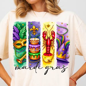 May include: A white t-shirt with a colorful graphic design featuring Mardi Gras symbols. The design includes a purple, green, and gold mask, a king cake, a red crawfish, and a purple, green, and gold jester hat. The text "Mardi Gras" is printed below the design.