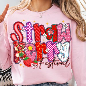 May include: A pale pink sweatshirt with the words "Strawberry Festival" in a colourful, whimsical design. The text features strawberries, stars, and flowers. The sweatshirt is a crew neck style.