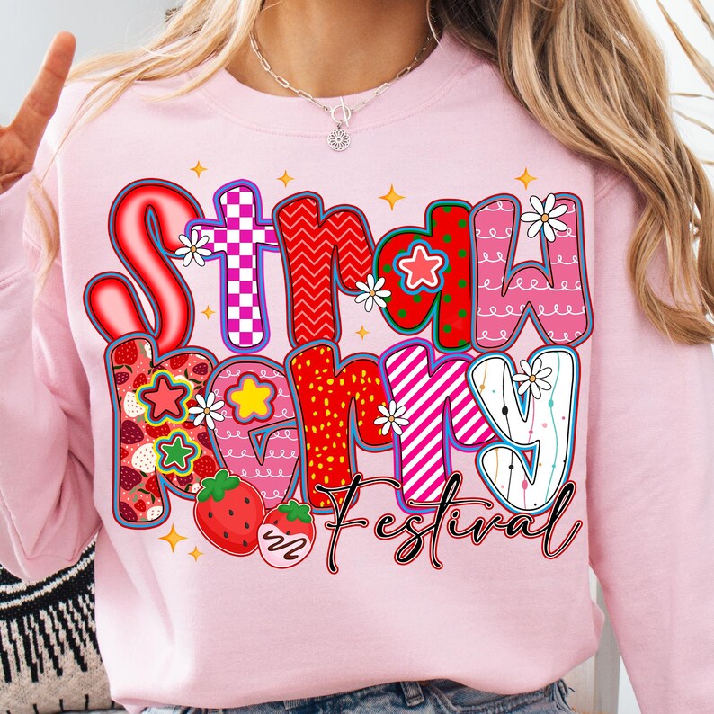 May include: A pale pink sweatshirt with the words "Strawberry Festival" in a colourful, whimsical design. The text features strawberries, stars, and flowers. The sweatshirt is a crew neck style.
