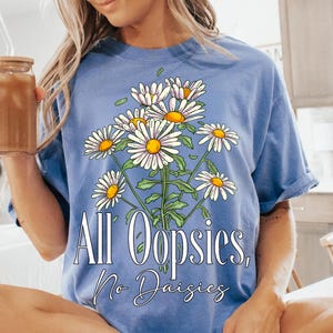 K&ouml;nnte beinhalten: Hellblaues T-Shirt mit einem G&auml;nsebl&uuml;mchen-Bouquet-Design. Die G&auml;nsebl&uuml;mchen haben wei&szlig;e Bl&uuml;tenbl&auml;tter und gelbe Zentren. Der Text "All Oopsies, No Daisies" ist aufgedruckt. Das T-Shirt hat einen l&auml;ssigen Stil.