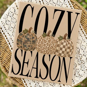 May include: A rectangular sign with a tan background featuring the black text COZY SEASON. Three pumpkins are centered between the words, each with a different pattern: one with green camouflage, one with leopard print, and one with a brown checkerboard.
