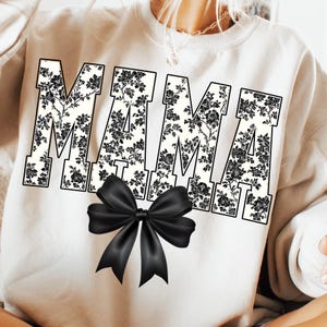 May include: Cream-colored sweatshirt with the word "MAMA" in large, block letters. The letters are filled with a black floral pattern. A large, black satin bow is centered below the text, adding a decorative touch to the design.