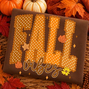 May include: A brown sweatshirt featuring the text FALL VIBES. The word FALL is written in large, orange, white-polka-dotted block letters with a lace-like border. The word vibes is written in a brown cursive script below, with small maple leaves and a yellow flower.