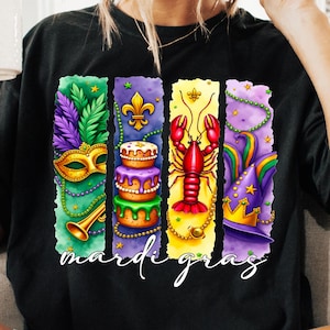 May include: A black t-shirt with a colorful design featuring Mardi Gras symbols. The design includes a mask, a cake, a crawfish, and a jester's hat. The text "Mardi Gras" is written below the design.