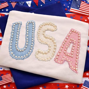 K&ouml;nnte beinhalten: Ein wei&szlig;es T-Shirt mit dem Wort "USA", das in Blau, Creme und Rosa gestickt ist, wobei jeder Buchstabe mit kleinen, wei&szlig;en Perlen verziert ist. Das Shirt wird vor einem Hintergrund patriotischer Dekorationen, einschlie&szlig;lich amerikanischer Flaggen, pr&auml;sentiert.