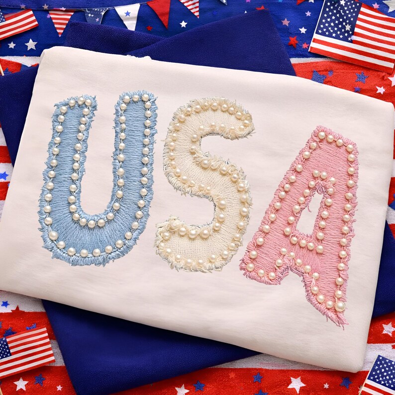 K&ouml;nnte beinhalten: Ein wei&szlig;es T-Shirt mit dem Wort "USA", das in Blau, Creme und Rosa gestickt ist, wobei jeder Buchstabe mit kleinen, wei&szlig;en Perlen verziert ist. Das Shirt wird vor einem Hintergrund patriotischer Dekorationen, einschlie&szlig;lich amerikanischer Flaggen, pr&auml;sentiert.