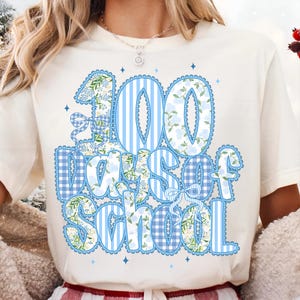 K&ouml;nnte beinhalten: Cremefarbenes T-Shirt mit dem Text "100 Days of School" in Blau und Wei&szlig;. Die Zahlen und Buchstaben haben Blumen- und Streifenmuster. Das Design umfasst Schleifen und kleine blaue Sternakzente.