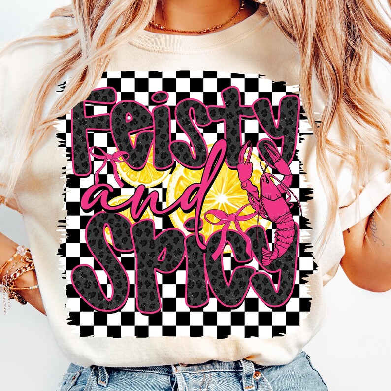 May include: A white t-shirt with a black and white checkered background. The text "Feisty and Spicy" is written in black with a pink leopard print pattern. There is a pink crawfish and lemons on the shirt.