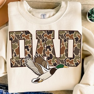 May include: Cream-colored sweatshirt with the word "DAD" in large, block letters filled with a camouflage pattern. A detailed illustration of a flying duck is beneath the text, featuring brown, white, green, and yellow colors.