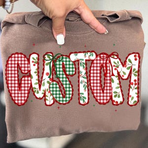 Pu&ograve; includere: Una felpa marrone con la scritta "CUSTOM" in un design festivo. Le lettere sono decorate con quadretti rossi e bianchi, quadretti verdi e bianchi con bacche rosse e foglie verdi. Fiocchi verdi e rossi accentuano le lettere.