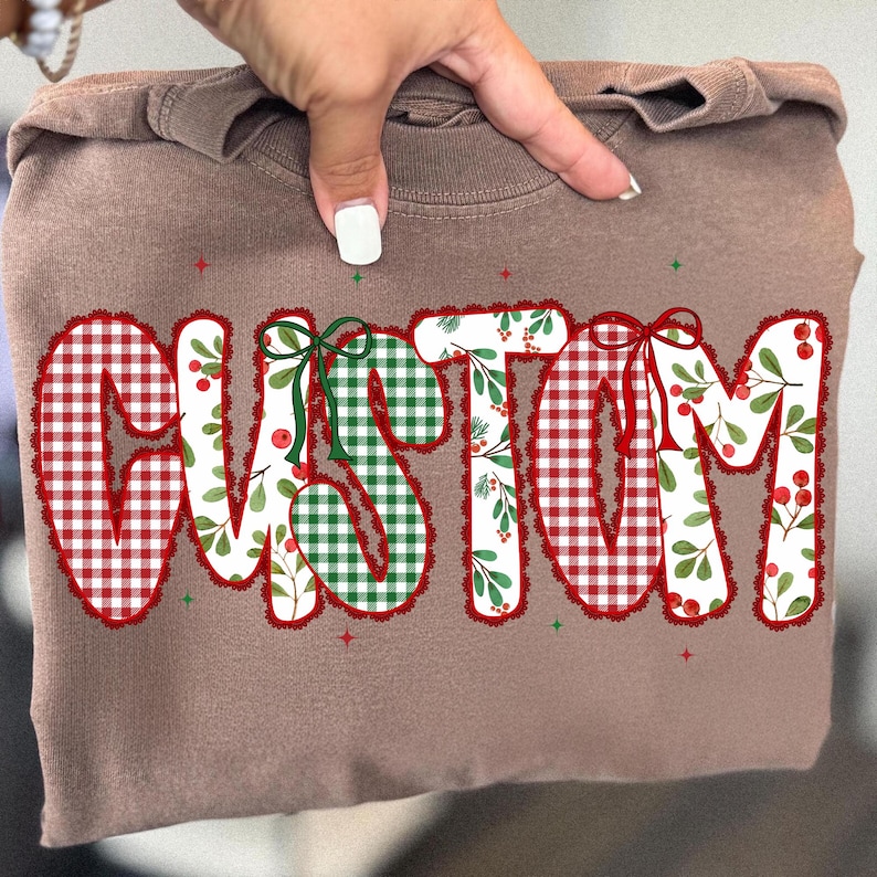 Pu&ograve; includere: Una felpa marrone con la scritta "CUSTOM" in un design festivo. Le lettere sono decorate con quadretti rossi e bianchi, quadretti verdi e bianchi con bacche rosse e foglie verdi. Fiocchi verdi e rossi accentuano le lettere.