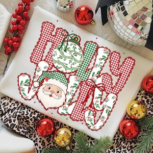May include: A cream-colored sweatshirt with the word "JOY" in a festive design. The letters are a mix of red gingham, green plaid, and a floral pattern. A Santa Claus graphic and Christmas ornaments complete the holiday theme.