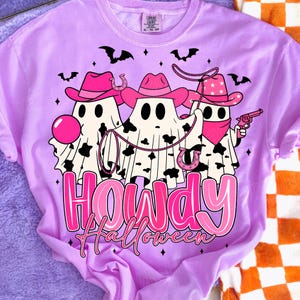 May include: A light purple t-shirt featuring a graphic of three ghost figures dressed as cowboys, wearing pink hats and bandanas. The ghosts have black and white cow-print patterns. The text "Howdy Halloween" is displayed in pink.