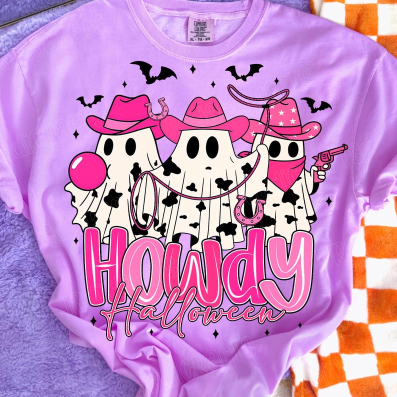 May include: A light purple t-shirt featuring a graphic of three ghost figures dressed as cowboys, wearing pink hats and bandanas. The ghosts have black and white cow-print patterns. The text "Howdy Halloween" is displayed in pink.