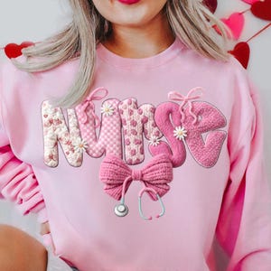 May include: A light pink sweatshirt with the word "NURSE" in a 3D design. The letters are decorated with floral and patterned details. A large pink bow and a stethoscope are below the word. The sweatshirt is a Valentine's Day theme.