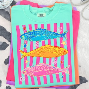 May include: A turquoise Comfort Colors t-shirt featuring a graphic of three colorful fish. The design includes vertical pink stripes, stars, and flowers. The fish are blue, yellow, and pink. The shirt is folded on top of other brightly colored shirts.