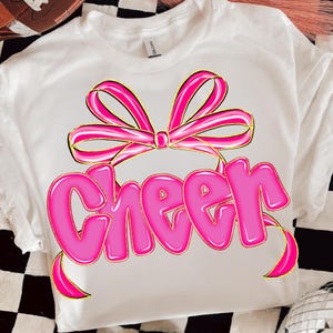 Puede incluir: Camiseta blanca con un lazo rosa grande y la palabra "Cheer" en letras de burbujas. El dise&ntilde;o tiene un contorno dorado, creando un efecto 3D. La camiseta est&aacute; hecha de un material suave.