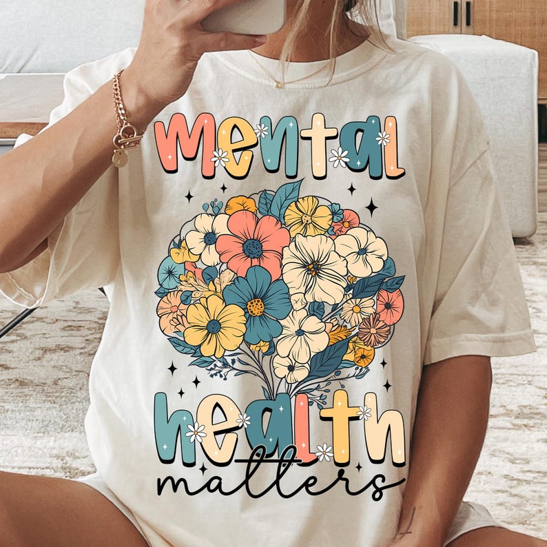 May include: Cream-colored t-shirt with the words "Mental Health Matters" in a colorful, floral design. The design features a bouquet of flowers in shades of pink, blue, yellow, and cream.
