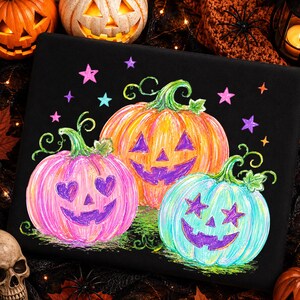 May include: A black fabric surface featuring a colorful illustration of three jack-o'-lanterns. The left pumpkin is pink with heart-shaped eyes, the center is orange, and the right is light blue with star-shaped eyes, all with purple faces.